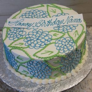 BR-029 is a round cake with blue and green floral patterns, piped cream borders, and a white plaque on top that says “Happy Birthday Nana” in blue script. The cake is presented on a foil-covered board.