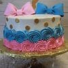 The BB-052 cake is round with gold polka dots, pink and blue bows, a gold question mark topper, and pink & blue rosette icing on the sides for a gender reveal theme, presented on a decorated stand.