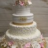 The WD-068 is a three-tiered white wedding cake with gold accents, intricate icing swirls, and large white, pink, and beige sugar flowers on a gold base set against a wooden background.