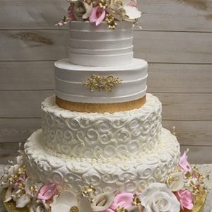 The WD-068 is a three-tiered white wedding cake with gold accents, intricate icing swirls, and large white, pink, and beige sugar flowers on a gold base set against a wooden background.