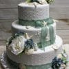 WD-080: Three-tier white cake with light green lace ribbon, adorned with white seashells, starfish, white roses, and blue hydrangeas on a foil-covered board against a wooden background.