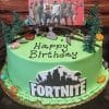 The CB-011D is a round green birthday cake topped with toy trees, candy rocks, tools, a "FORTNITE" logo, and "Happy Birthday" lettering. It features a topper displaying Fortnite characters in the background.
