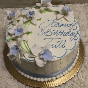 The BD-104 cake is a round white cake with blue and white floral decorations, blue piped dots, and "Happy Birthday Tutti" written in blue cursive icing on top. It is presented on a gold scalloped cake board.