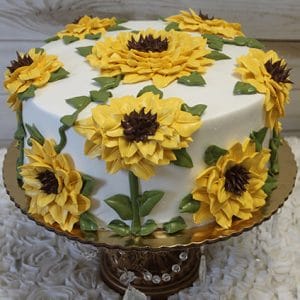 BR-055 is a round white cake adorned with large yellow sunflower icing designs, green leaves, and brown centers, presented on a gold cake stand.