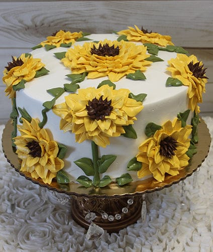 The THX-032 is a round cake with smooth white icing, decorated with large yellow sunflowers and green leaves. It sits on a gold stand with hanging crystals, set against a light wooden background.