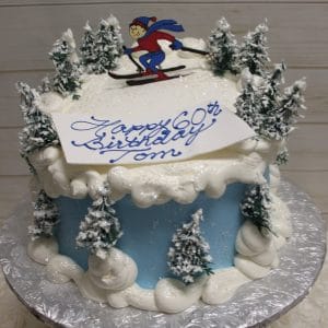 The ST-011 Skiing cake is round with blue and white icing, features snow-covered trees and a skier topper, and comes with a “Happy 60th Birthday, Tom” card in blue script on top. Presented on a silver cake board.