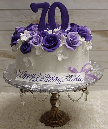 The BD-060H Shades of Purple Flowers cake is white, decorated with purple and white flowers, topped with a large purple 70, displayed on a stand, with a plaque reading "Happy Birthday Hilda" in script.