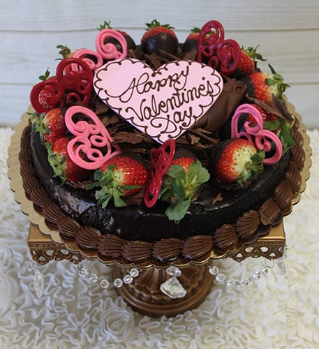 VC-010 is a chocolate cake topped with strawberries, pink heart decorations, and a heart-shaped plaque that says "Happy Valentines Day," presented on a decorative cake stand.