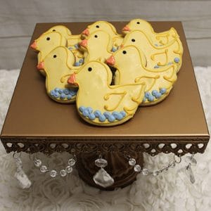 A set of CE-004e baby shower cookies—yellow duck shapes with orange beaks and blue icing—are displayed on a gold stand decorated with hanging crystal ornaments.