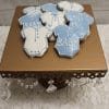 The CE-004a baby shower features blue and white iced cookies shaped like baby onesies, some with "BABY" written on them, presented on a gold cake stand with crystal accents atop a white textured tablecloth.