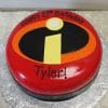 CB-006F is a round, red cake featuring The Incredibles logo and "Happy 5th Birthday Tyler!" in black icing, presented on a silver floral-patterned board.