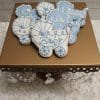 The CE-004c baby shower features sugar cookies shaped like carriages with blue and white icing, some labeled "Baby," displayed on a gold stand with crystal accents atop a white floral surface.