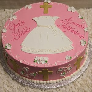 The RC-036 is a round pink cake with a white dress, gold crosses, white flowers, and "God Bless Anna Katherine" in pink icing, displayed on a white ruffled tablecloth.