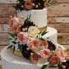 WD-086: A three-tiered white cake with gold ribbon accents, adorned with pink, peach, and burgundy flowers, green leaves, and gold berries, set against a rustic wooden background.