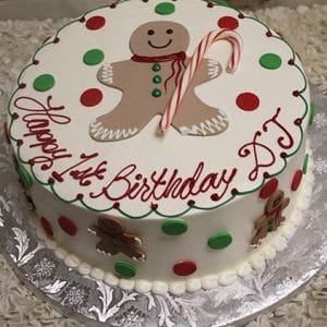CH-027: Round cake topped with red and green polka dots, a smiling gingerbread man, and a candy cane. Sides feature small gingerbread men and “Happy 1st Birthday DJ!” written in red icing.