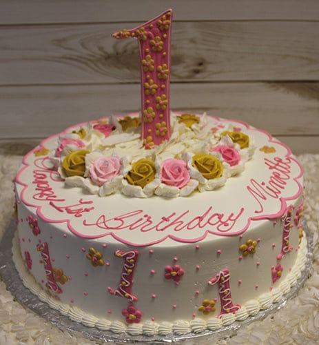 CB-023C: Round white cake with pink and gold flowers, pink and gold number ones, topped with a large pink number one and roses. “Happy 1st Birthday Nimette” is written in pink icing.