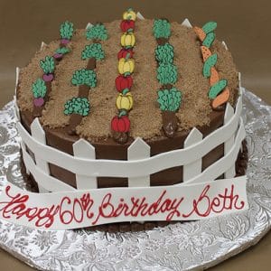 BD-081 is a round cake designed as a vegetable garden with rows of icing vegetables, a white picket fence, and “Happy 60th Birthday Beth” written in red on a banner.