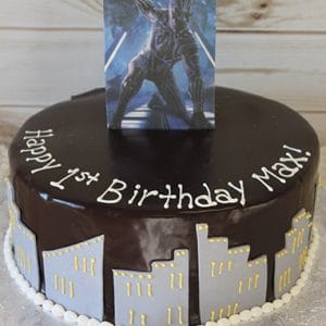The CB-006G is a chocolate-glazed birthday cake with a city skyline, white and yellow icing, a superhero card, and “Happy 1st Birthday Max!” piped in white icing.