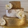 The WD-087 is a three-tiered cake with gold and white layers, adorned with gold sprinkles and large white sugar flowers, displayed on a gold embossed base against a wooden background.