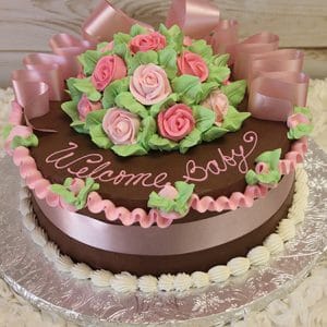 The BB-014 cake features pink and green icing roses, pink ribbon bows, and “Welcome Baby” in pink icing on top. It’s presented on a silver-covered board with a white frosting border.