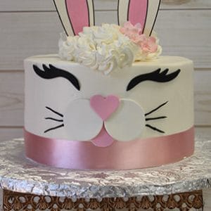 The EA-022 cake features a round bunny face with tall fondant ears, closed eyes, a pink heart-shaped nose, whiskers, and white and pink flowers on top, displayed on a silver stand with crystal accents.