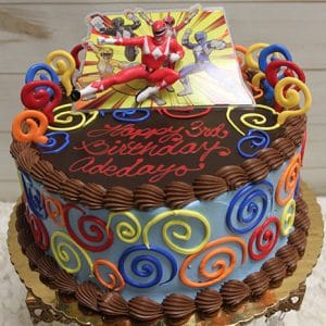 A festive SC-058 Power Rangers birthday cake with chocolate swirls, red, yellow, and blue icing, topped with Power Rangers figures and “Happy 3rd Birthday Adebayo” written in red icing.