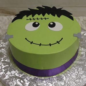 The HW-009 is a round cake designed as a cartoon Frankenstein’s monster with green icing, black hair, big eyes, a stitched forehead scar, and a purple ribbon around the base, set on a silver floral-patterned board.