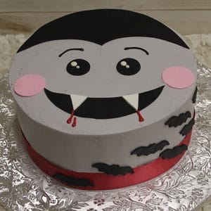 The HW-008 is a round cake designed as a cartoon vampire face with fangs, pink cheeks, and dripping blood. Icing bats decorate the sides, a red ribbon trims the base, and it rests on a silver-patterned board.