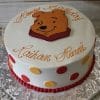 CB-009P is a round white birthday cake featuring red and yellow polka dots, a red bead border, Winnie the Pooh’s face on top, and “Happy Birthday Raihan Hunni” written in orange script.