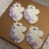Four CE-001e birthday cookies shaped like unicorns, with white icing, purple and pink manes, and closed eyes with long lashes, are displayed on a gold rectangular tray.