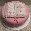 RC-095 is a round pink cake designed as an open book, with white icing, a pink cross, and "God Bless Cailyn" in cursive. It rests on a silver board and is accented by white flowers.