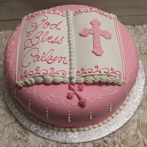 RC-095 is a round pink cake designed as an open book, with white icing, a pink cross, and "God Bless Cailyn" in cursive. It rests on a silver board and is accented by white flowers.