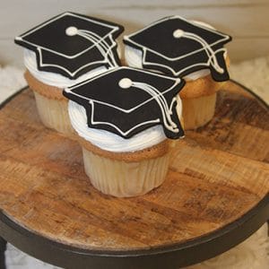Three CC-035 Gold cupcakes with chocolate filling are displayed on a round wooden stand, each topped with white frosting and black graduation cap-shaped cookies.