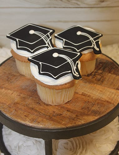 Three GC-072 gold cupcakes with chocolate filling are topped with black graduation cap icing decorations and displayed on a wooden cake stand.