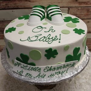 BB-026: Round cake with green polka dots and shamrocks, fondant baby shoes topper, “Oh Baby!” iced on top, and side banner reading “A Little Shamrock is on his way!”.