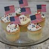 Six JC-021 chocolate cupcakes with white chocolate mousse filling, topped with white frosting, red and blue star sprinkles, and small American flags, are arranged on a glass cake stand against a light wooden background.