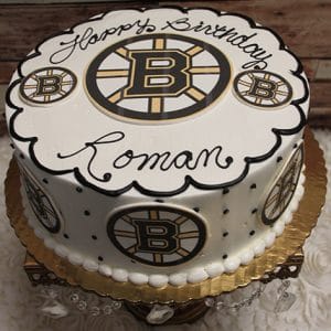 A round ST-006 Bruins cake features the Boston Bruins logo, black and gold accents, “Happy Birthday Roman” in black icing, and is presented on a gold board.
