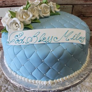 RC-066 is a round, light blue cake featuring a quilted pattern with white pearl accents, topped with white icing roses and green leaves. A plaque on top reads "God Bless Miles" in blue script.