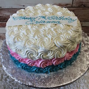 CB-018A is a round cake with white, pink, and blue swirled icing. A white plaque on top says “Happy 7th Birthday Arianna” in blue script. The cake is presented on a silver, textured board.