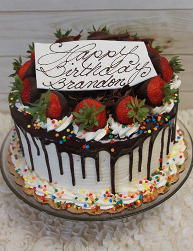 The BD-007 is a chocolate-dripped birthday cake with strawberries, whipped cream, and colorful sprinkles. A white plaque says “Happy Birthday Brandon” in cursive. The cake sits on a glass stand against a light wood background.