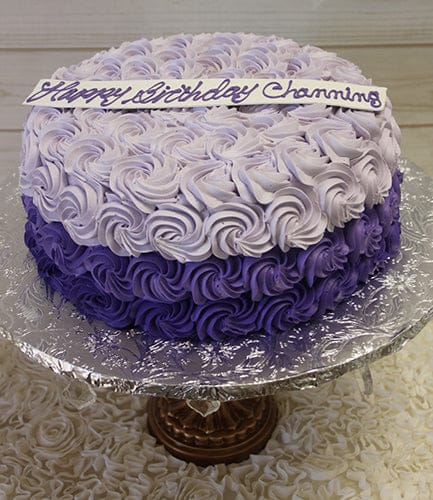 The CB-018C is a round cake with light and dark purple rosette frosting on a silver stand, topped with a white plaque that says "Happy Birthday Channing" in purple script.