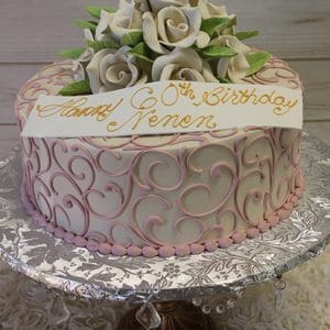 The BD-060C Ivory Roses cake features pink swirl decorations, white fondant roses with green leaves, and a white banner with gold "Happy 60th Birthday Nanon" lettering, presented on an ornate silver stand.
