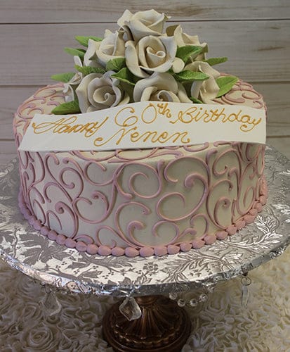 The BD-060C Ivory Roses cake features pink swirl decorations, white fondant roses with green leaves, and a white banner with gold "Happy 60th Birthday Nanon" lettering, presented on an ornate silver stand.