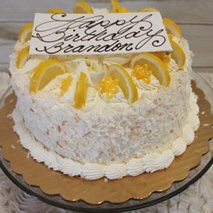 The BD-004 Lemon Citron is a round, white frosted cake topped with lemon slices, yellow flowers, and a rectangular plaque reading "Happy Birthday Brandon," all presented on a gold platter.