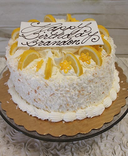 The BD-004 Lemon Citron is a round, white frosted cake topped with lemon slices, yellow flowers, and a rectangular plaque reading "Happy Birthday Brandon," all presented on a gold platter.