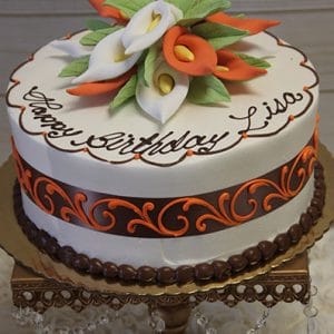 BD-060L White/Orange Calla Lillies: A round white birthday cake with "Happy Birthday Lisa" in brown icing, topped with orange and white calla lilies, orange scroll design, and a dark brown ribbon around the sides.