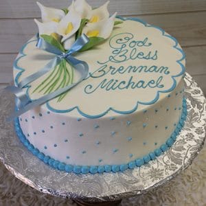 RC-010: Round white cake with blue polka dots and border, topped with white calla lilies, a blue ribbon, and "God Bless Brennan Michael" in blue icing. Presented on a silver-patterned board.