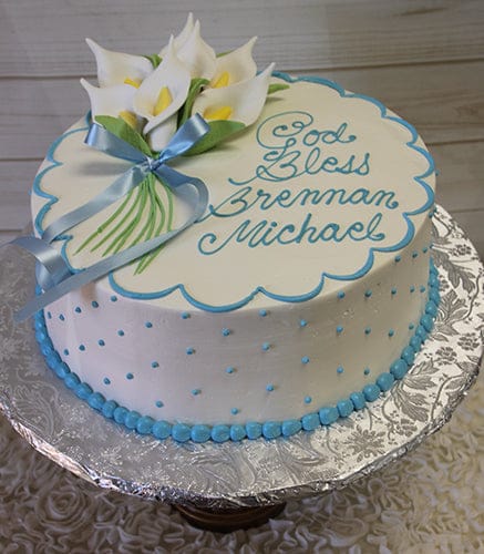 RC-010: Round white cake with blue polka dots and border, topped with white calla lilies, a blue ribbon, and "God Bless Brennan Michael" in blue icing. Presented on a silver-patterned board.
