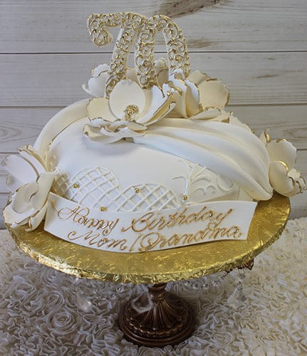 The BD-055C White Swag is a white and gold birthday cake featuring decorative flowers, gold accents, and a "70" topper with a “Happy Birthday Mom Grandma” plaque, elegantly displayed on a gold stand against a wooden background.