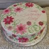 The BD-060 Shades of Pink Daisies cake features pink and red fondant flowers, green swirls, and “Happy Birthday Mary” in pink icing. It’s displayed on a silver board with white ruffled fabric beneath.